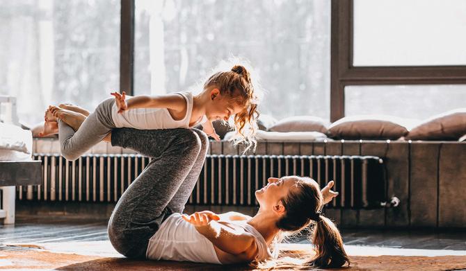 Mutter liegt mit junger Tochter auf dem Fußboden und hat diese auf den Beinen abgestemmt. Im Hintergrund ist ein durch Fernwärme betriebener Heizkörper zu sehen.