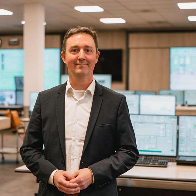 Freundlich lächelnder Mann im Anzug steht in einem Raum mit vielen Monitoren, auf denen Schaltpläne zu erkennen sind.
