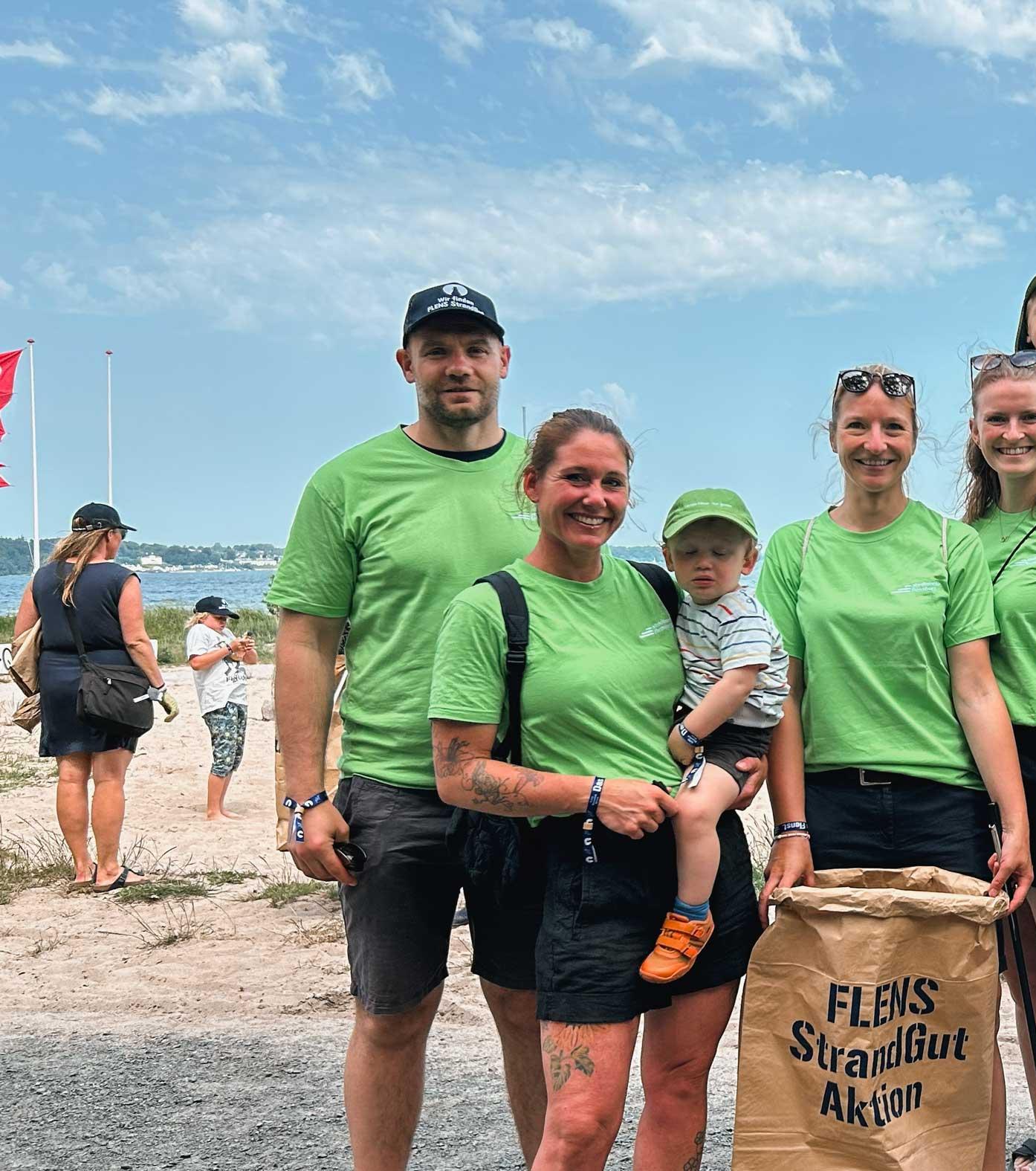Eine Gruppe von Freiwilligen, Erwachsenen und Kinder, lächelt am Strand nach einer Strandreinigungsaktion. Die Teilnehmer tragen grüne T-Shirts und halten braune Papiertüten mit dem Aufdruck "FLENS StrandGut Aktion". Im Hintergrund ist die Flensburger Förde zu sehen.
