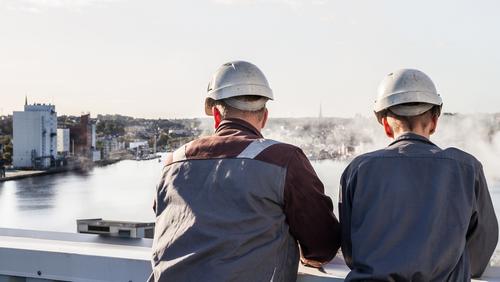 2 Handwerker mit Schutzhelmen stehen auf dem Dach der Stadtwerke Flensburg und schauen über die FLensburger Förde.