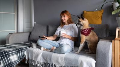Junge Frau sitzt mit einem Hund auf einem Sofa, in der Hand hält sie eine Fernbedienung in der anderen Hand einen Kaffeebecher
