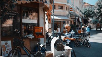 Älterer Herr mit Brille und T-Shirt steht an einer Straßenecke in einer türkischen Stadt vor einem Laden im Sonnenschein