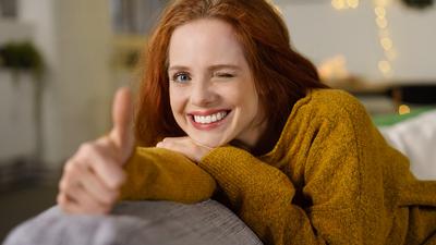 Eine junge Frau mit rötlichen Haaren und einem gelbem Pulli lehnt mit dem Oberkörper auf einem Sofa. Sie lächelt, zwinkert und hat ihren Daumen aufgerichtet. Im Hintergrund ist eine Lichterkette zu erkennen.