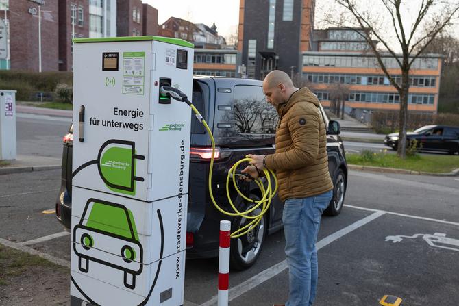 Mann im öffentlichen Raum steht vor E-Ladesäule mit Stadtwerke Flensburg Logo und Icon und verbindet schwarzes Auto mit gelbem Kabel.