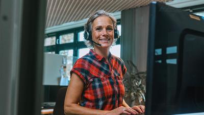 Lächelnde Frau steht mit einem Headset an einem Schreibtisch mit Monitor und trägt eine karierte Kurzarmbluse.