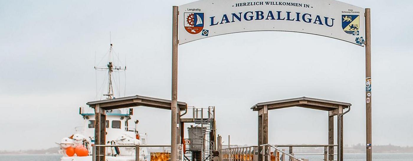 Brücke mit der Aufschrift Langballigau. Linksseitig hat ein Schiff an der Brücke angelegt. Im Hintergrund ist das Wasser und eine Landzunge zu sehen.
