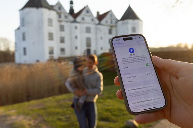 Eine Frau mit Kind auf dem Arm steht vor einem weißen Schloss. Im Vordergrund hält eine Hand ein Handy mit WLAN-Verbindung im Display.