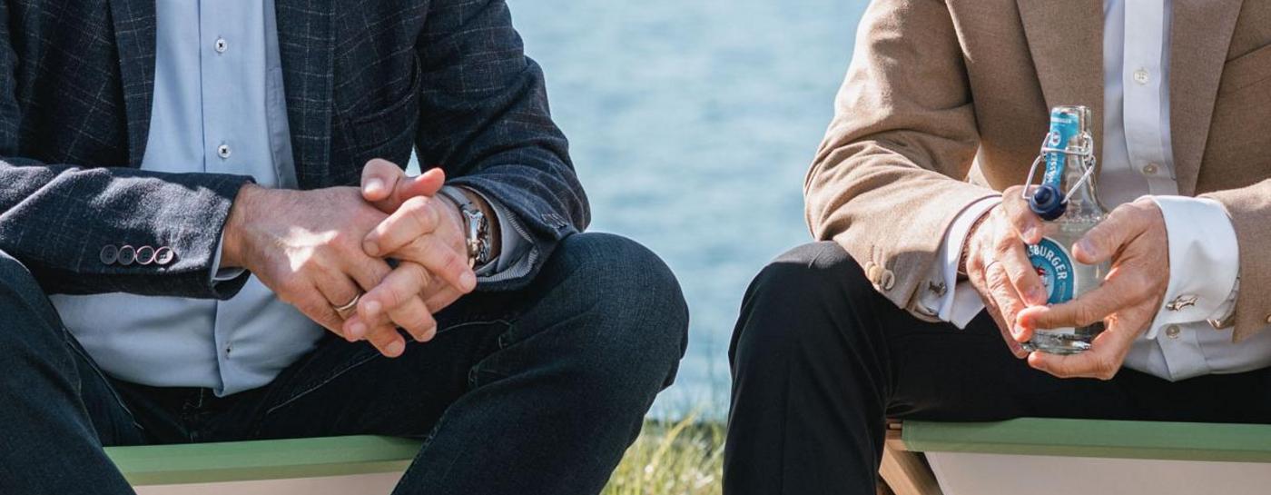 Zwei lächelnde Männer in eleganten Sakkos sitzen nebeneinander in Liegestühlen an einer Uferpromenade vor einer sommerlichen Meereskulisse.