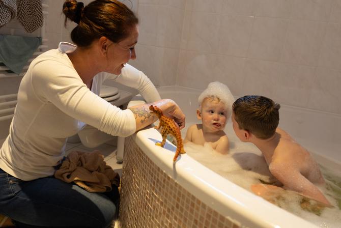 Zwei Kinder sitzen in einer gefüllten Badewanne. Eine junge Frau kniet davor mit Blick auf die Kinder. Das jüngere Kind hat Schaum auf dem Kopf.