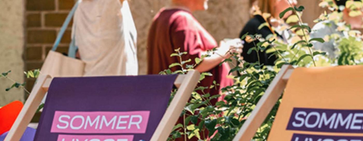 Zwei Liegestühle mit dem Logo „Sommer Hygge Flensburg“ in einem grün bepflanzten Innenhof mit bunten Wimpelketten und Besuchern im Hintergrund