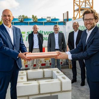 Zwei Männer in Anzügen halten lachend ein kupferfarbenenes Rohr über einen Steinschacht. Im Hintergrund stehen drei weitere Männer.