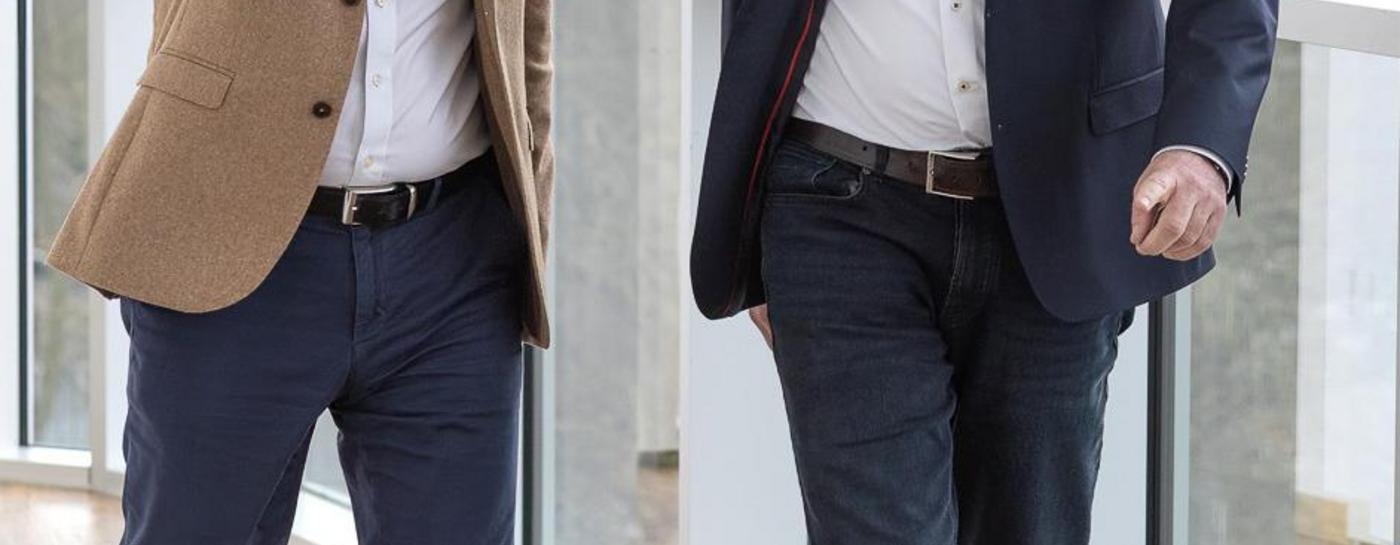 Zwei Männer gehen nebeneinander einen Holzflur an einer großen Fensterfront entlang. Dabei schauen sie sich an. Der Mann links gestikuliert mit seinem rechten Arm und Hand. Beide tragen Hemd und Sakko.