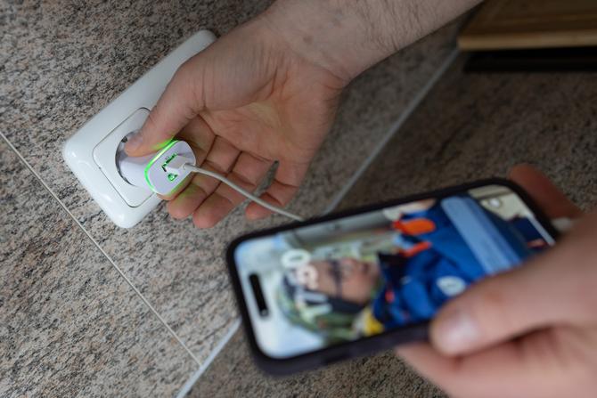 Der Stecker eines Handyladekabels wird in die Steckdose gedrückt. Das Handy zeigt einen Jungen auf dem Hintergrundbild.