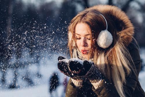 Junge Frau mit Winterjacke, Fellkragen und Ohrschützern pustet Schnee von ihren behandschuhten Händen