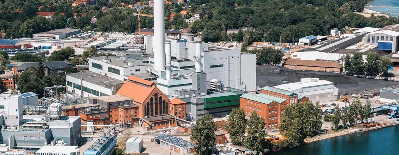 Luftaufnahme des Stadtwerke Flensburg Betriebsgeländes an der Förde mit Kraftwerk, hohem Schornstein, Hafenanlagen und Werftgebäuden. Im Hintergrund ist ein Wald und eine Wohnsiedlung zu sehen.