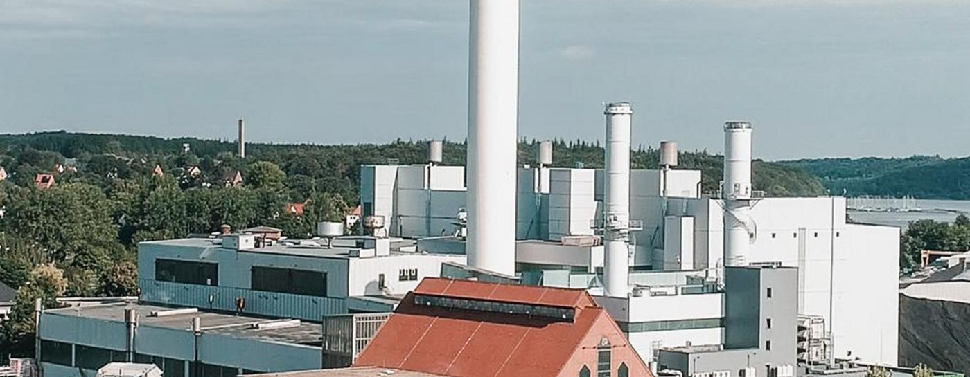 Stadtwerke Gelände aus der Vogelperspektive bei blauem Himmel mit einigen Wolken. Im Hintergrund sind die Flensburger Förde und Bäume zu sehen.