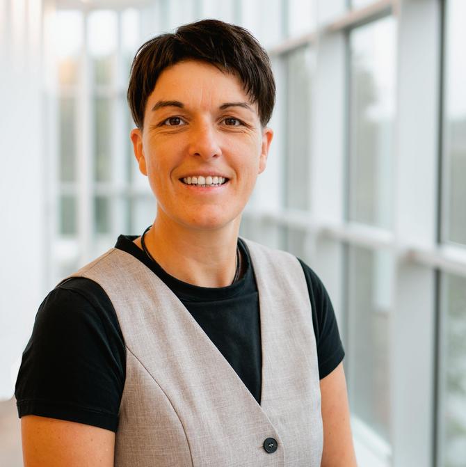 Portrait einer lächelnden Frau mit dunklen, kurzen Haaren. Sie steht vor einer Fensterfront und trägt ein schwarzes T-Shirt mit beiger Weste.