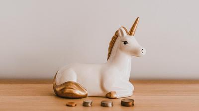 Weißes Einhorn liegt hinter einer Reihe gestapelter Cent-Münzen-Haufen auf einem Holzboden, weißer Hintergrund.