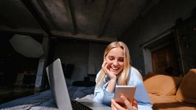 Frau liegt mit Laptop und Handy auf dem Bett und nutzt das schnelle Glasfaser-Internet der Stadtwerke Flensburg.