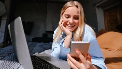 Frau liegt mit Laptop und Handy auf dem Bett und nutzt das schnelle Glasfaser-Internet der Stadtwerke Flensburg.