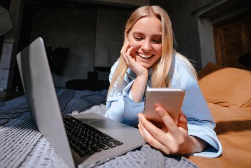 Frau liegt mit Laptop und Handy auf dem Bett und nutzt das schnelle Glasfaser-Internet der Stadtwerke Flensburg.