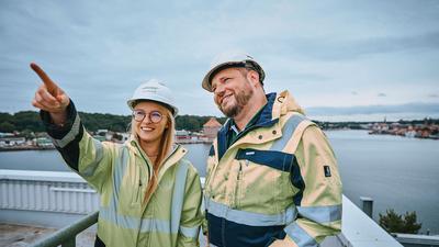 Eine blonde junge Frau und ein junger, großer Mann stehen auf einen hohen Gebäude und tragen Stadtwerke Kleidung und Helm. Beide schauen lächelnd. Die Frau zeigt mit dem Zeigefinger in Richtung Kamera. Im Hintergrund ist Wasser und zwei Landseiten mit Häusern zu sehen.
