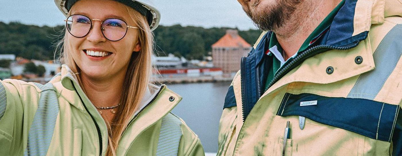 Eine blonde junge Frau und ein junger, großer Mann stehen auf einen hohen Gebäude und tragen Stadtwerke Kleidung und Helm. Beide schauen lächelnd. Die Frau zeigt mit dem Zeigefinger in Richtung Kamera. Im Hintergrund ist Wasser und zwei Landseiten mit Häusern zu sehen.