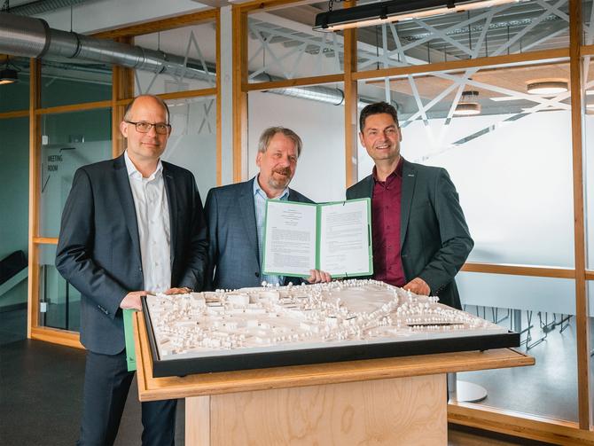 Drei lächelnde Männer in Anzug stehen vor einem Stadtmodell als Platte. Der mittlere Mann hat eine geöffnete Mappe mit zwei bedruckten Blättern in der Hand. Im Hintergrund ist ein Glasbüro zu erkennen.