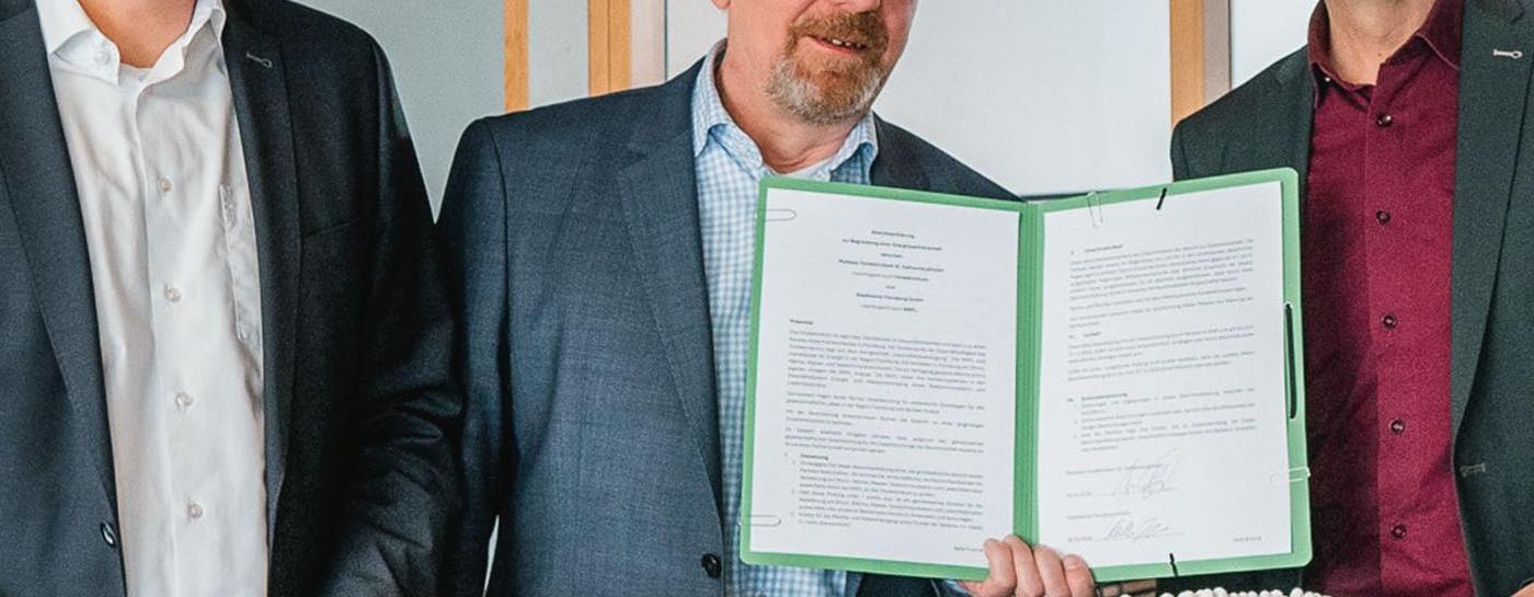 Drei lächelnde Männer in Anzug stehen vor einem Stadtmodell als Platte. Der mittlere Mann hat eine geöffnete Mappe mit zwei bedruckten Blättern in der Hand. Im Hintergrund ist ein Glasbüro zu erkennen.