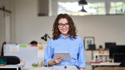 Eine Junge Frau mit Locken und einer Blau-Weiß gestreiften Bluse steht im Büro und hält ein Ipad in der rechten Hand und grinst.