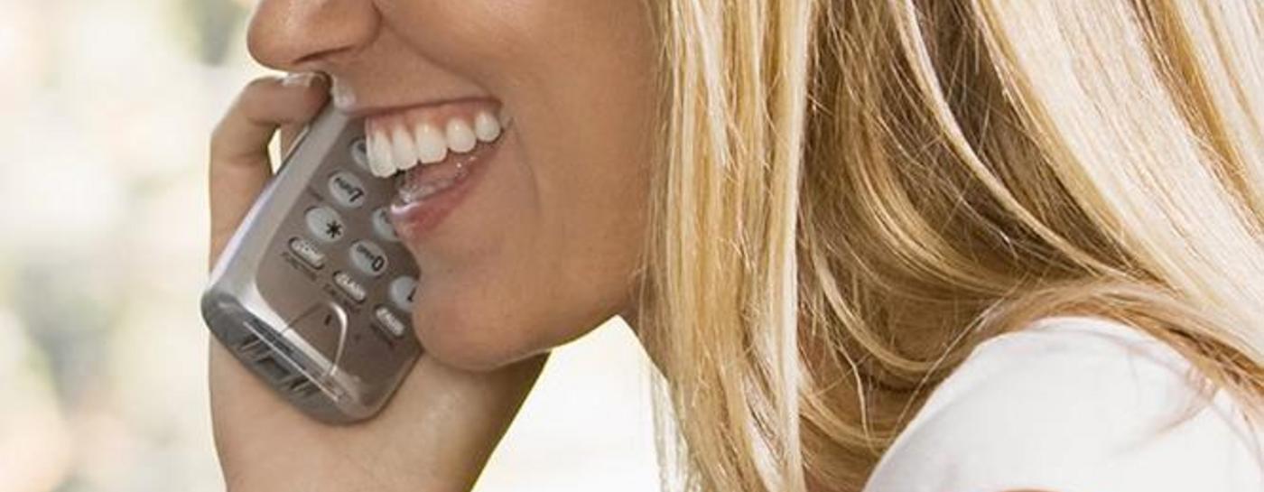 Eine blonde Frau mit langen Haaren sitzt lächelnd am Schreibtisch vor Ihrem Computer und telefoniert. Für Internet und Telefonie nutzt sie einen Glasfaser-Anschluss der Stadtwerke Flensburg.