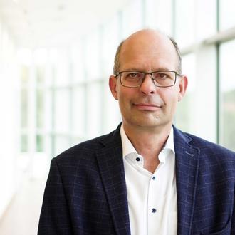 Portrait von Karsten Mueller-Janssen in Anzug und weißem Hemd. Im Hintergrund ist eine Fensterscheibenfront.