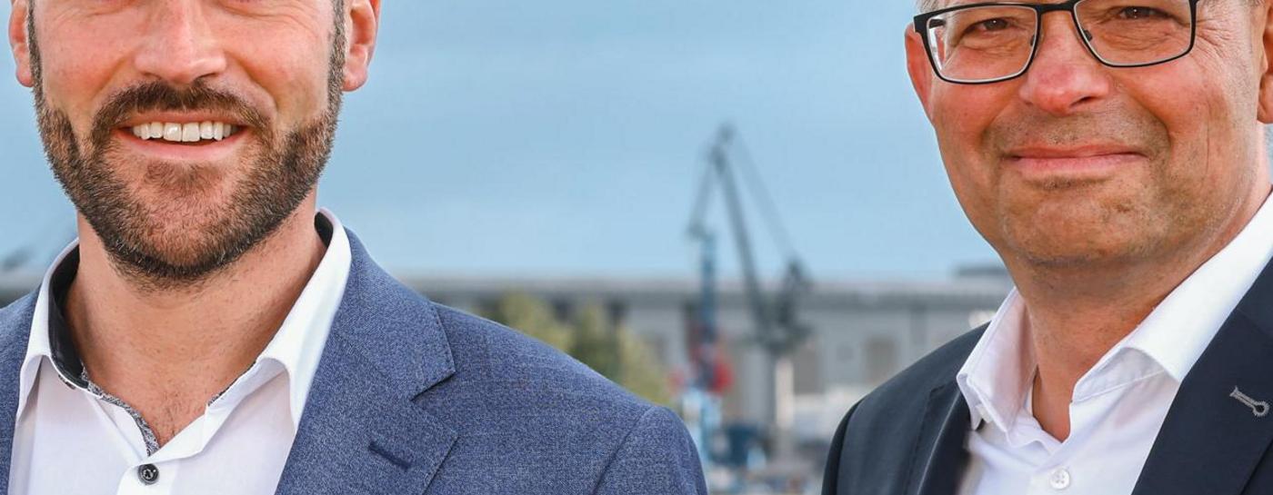 Die Geschäftsführer der Stadtwerke Flensburg, rechts Karsten Müller-Janßen, links Julian Wollscheidt, beide tragen ein weißes Hemd und Sakko, stehen draußen mit der Förde und dem Gelände der Stadtwerke Flensburg im Hintergrund und gucken freundlich in die Kamera.