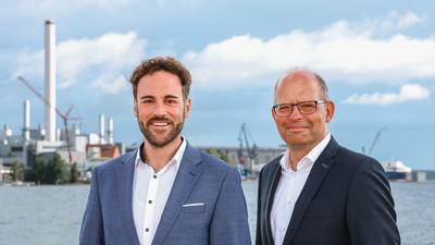 Die Geschäftsführer der Stadtwerke Flensburg, rechts Karsten Müller-Janßen, links Julian Wollscheidt, beide tragen ein weißes Hemd und Sakko, stehen draußen mit der Förde und dem Gelände der Stadtwerke Flensburg im Hintergrund und gucken freundlich in die Kamera.