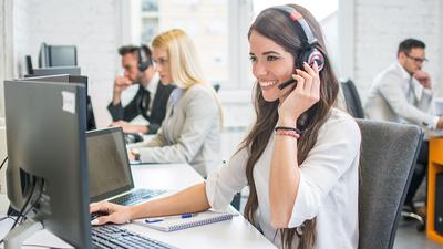 Eine junge Frau sitzt an einem Schreibtisch im Büro. Sie telefoniert mit einem Headset. Auf dem Schreibtisch sind 2 Monitore, 1 Laptop sowie Tastatur und ein Block mit einem Kugelschreiber.
