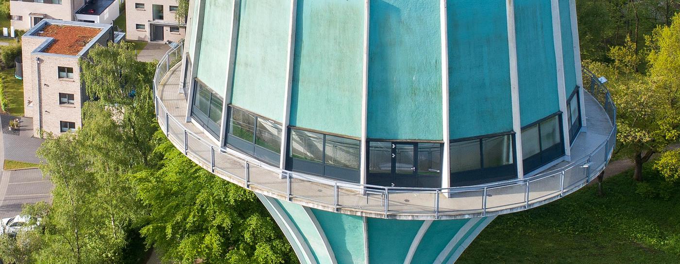 Blick von oben auf den hellgrünen Wasserturm Flensburg, der von Bäumen und modernen Mehrfamilienhäusern umgeben ist. 