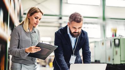 Eine Frau und ein Mann stehen zusammen im Lager. Die Frau hält eine Mappe in der Hand und schaut konzentriert, Rechts daneben steht der Mann welcher am Laptop arbeitet.
