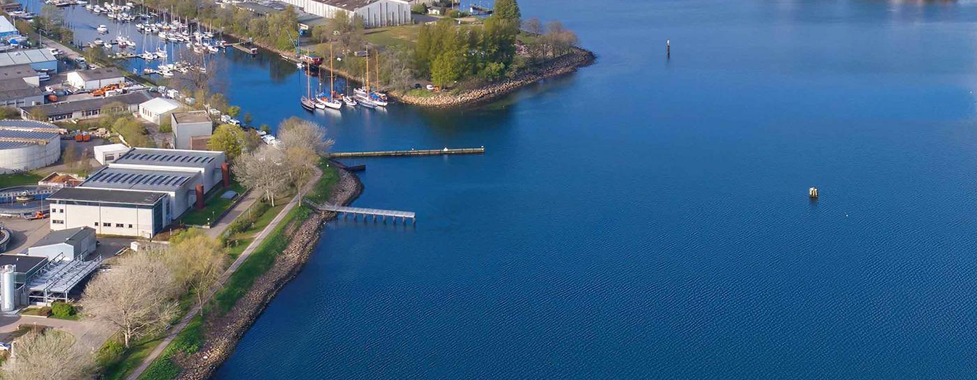 Flensburger Förde von oben bei blauem Himmel mit einigen Wolken. Linksseitig ist das Klärwerk zu sehen, rechts die Stadtwerke Flensburg und am Horizont die Spitze der Förde sowie Gebäude von Flensburg.
