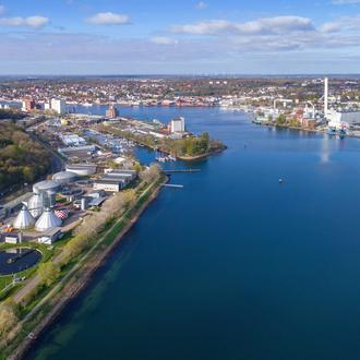 Flensburger Förde von oben bei blauem Himmel mit einigen Wolken. Linksseitig ist das Klärwerk zu sehen, rechts die Stadtwerke Flensburg und am Horizont die Spitze der Förde sowie Gebäude von Flensburg.