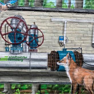 Bunt bemaltes Steinhäuschen mit Vogel, Fuchs und Rohren der Stadtwerke Flensburg.