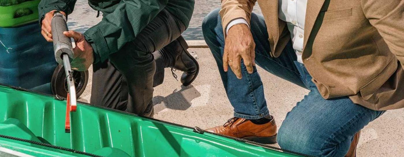 Drei freundlich schauende Männer knien vor einem grünen Kajak mit der Aufschrift GreenKayak. Im Hintergrund ist das Wasser und Häuser einer Stadt zu erkennen.