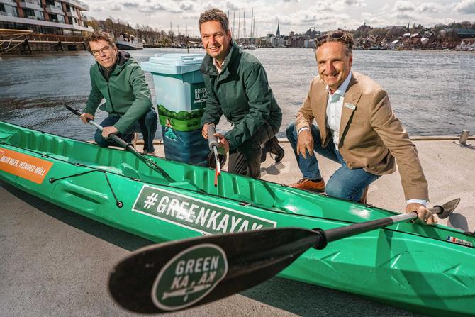 Drei freundlich schauende Männer knien vor einem grünen Kajak mit der Aufschrift GreenKayak. Im Hintergrund ist das Wasser und Häuser einer Stadt zu erkennen.