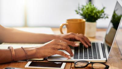 Ein Laptop mit Glasfaser-Anschluss der Stadtwerk Flensburg steht auf einem Holzschreibtisch. Eine weibliche Person tippt auf der Tastatur. Neben dem Laptop stehen ein Kaffeebecher und eine Pflanze. Eine Lesebrille liegt auf dem Tisch.