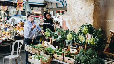 Mann mit Brille, Mütze und Zigarette im Mundwinkel steht an einem Marktstand in einer italienischen Stadt und preist Waren an