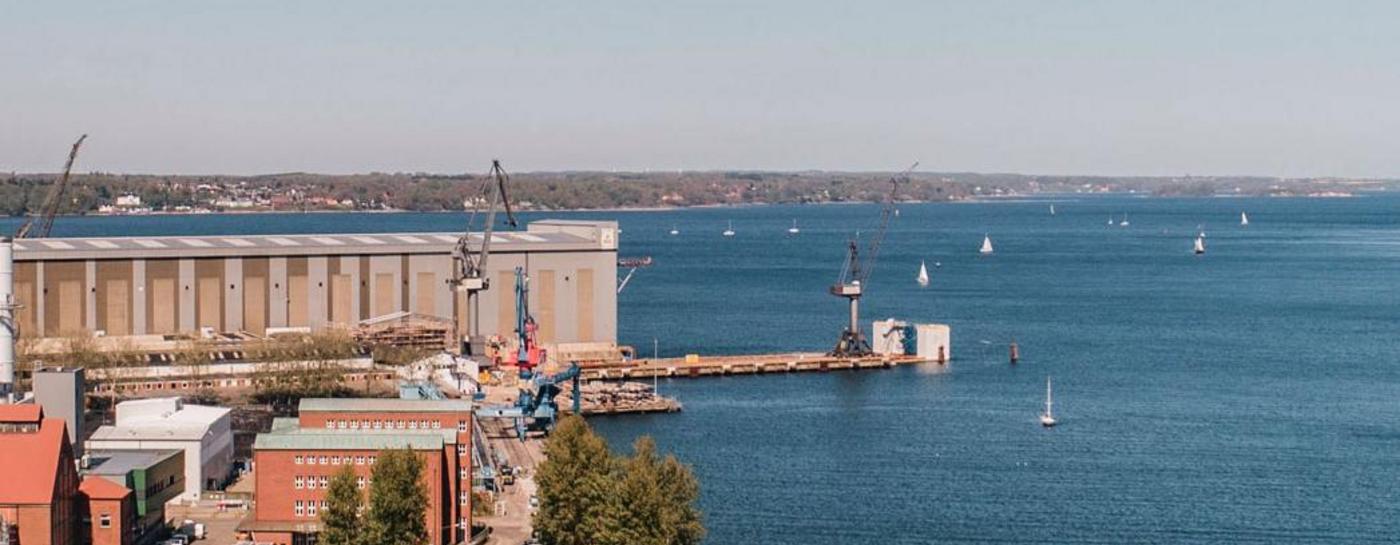 Gelände der Stadtwerke Flensburg von oben fotografiert bei Sonnenschein und klarem Himmel und ruhiger Förde. Auf dem Wasser sind Schiffe.