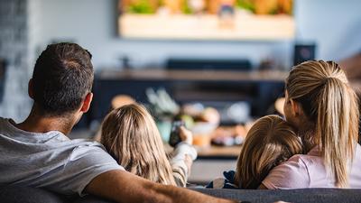 Eine Familie, die einen Glasfaser-Anschluss der Stadtwerke Flensburg nutzt, ist von hinten zu sehen. Sie sitzt auf einem Sofa und schaut Glasfaser-TV.