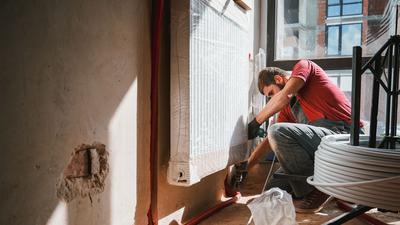 Sanierungsarbeiten im Inneren eines Raumes. An linken Wand ist ein kleiner Ausschnitt Mauerwerk freigelegt. Ein Heizungskörper wird durch einen Monteur angebracht. Im Hintergrund ist ein neues Fenster eingebaut.
