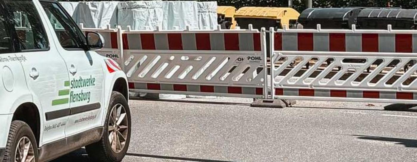 Weißes Stadtwerke Flensburg Baustellenfahrzeug vor einer abgesperrten Straße bei Sonnenschein und Bäumen im Hintergrund.