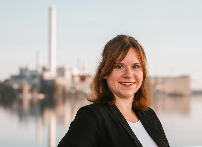 Junge Frau lächelt in die Kamera. Sie trägt ein weißes Shirt und dunklen Blazer. Im Hintergrund ist das Kraftwerk der Stadtwerke und die Flensburger Förde.