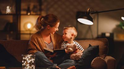 Eine Frau sitzt mit einem kleinen Jungen auf dem Sofa. Er hält einen Teddy, sie ein Buch, das Licht ist gedämpft.